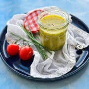 Angled view of classic French vinaigrette in a jar with fresh chives and cherry tomatoes on a dark blue plate.