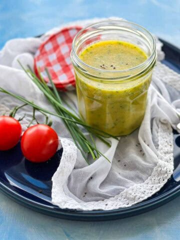 Angled view of classic French vinaigrette in a jar with fresh chives and cherry tomatoes on a dark blue plate.