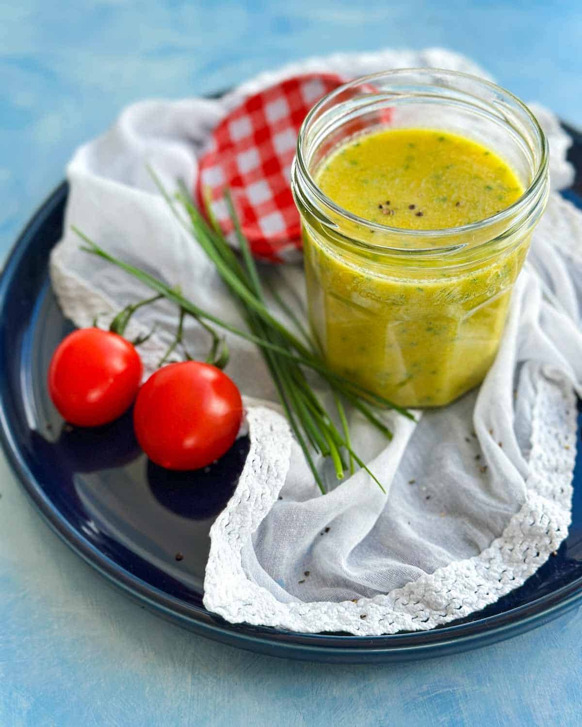 Angled close-up of classic French vinaigrette in a glass jar with fresh chives and cherry tomatoes on a blue plate.