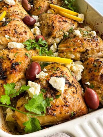 Angled close-up of Greek chicken and lemon potatoes in a baking dish with roasted chicken thighs, feta, olives, and parsley.