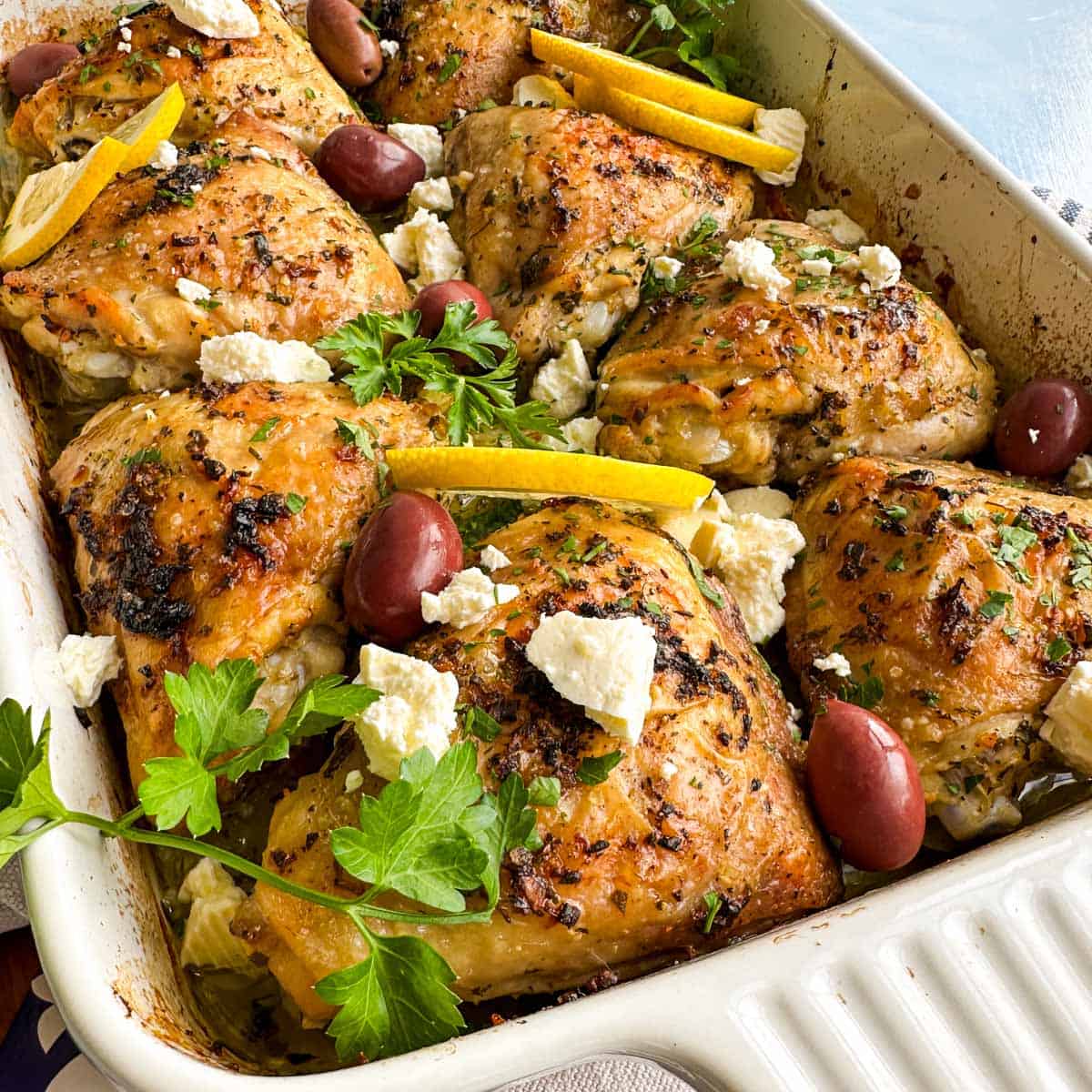 Angled close-up of Greek chicken and lemon potatoes in a baking dish with roasted chicken thighs, feta, olives, and parsley.