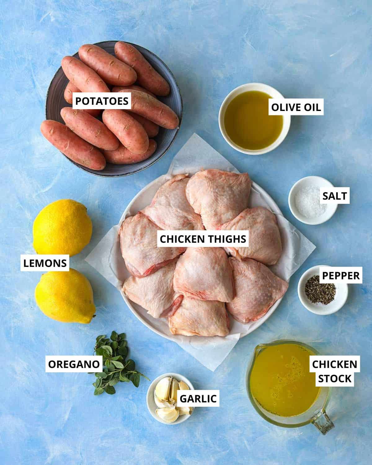 Top view of ingredients for Greek chicken and lemon potatoes including chicken thighs, potatoes, lemons, garlic, oregano, olive oil, stock, salt, and pepper.
