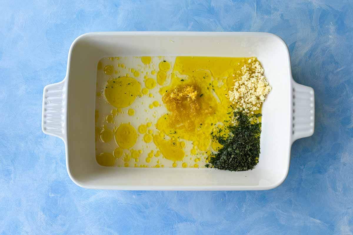 Top view of olive oil, lemon zest, garlic, and oregano in a baking dish for Greek chicken and lemon potatoes.