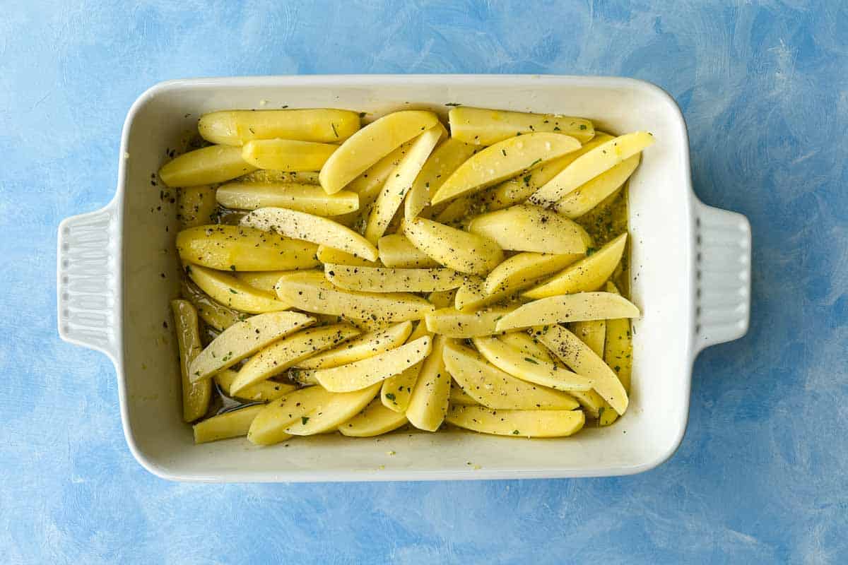 Top view of potato wedges in a baking dish with lemony herb marinade for Greek chicken and lemon potatoes.