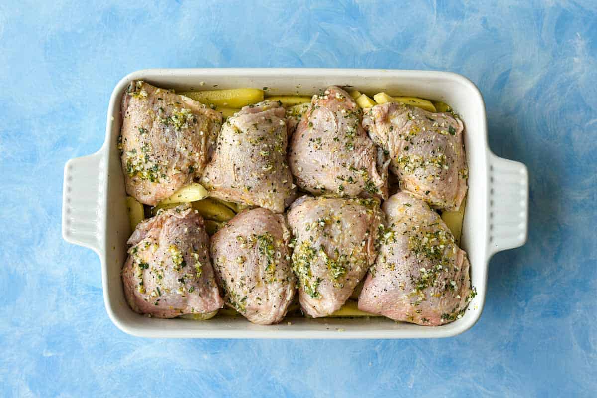 Top view of raw chicken thighs arranged over potato wedges in a baking dish for Greek chicken and lemon potatoes.