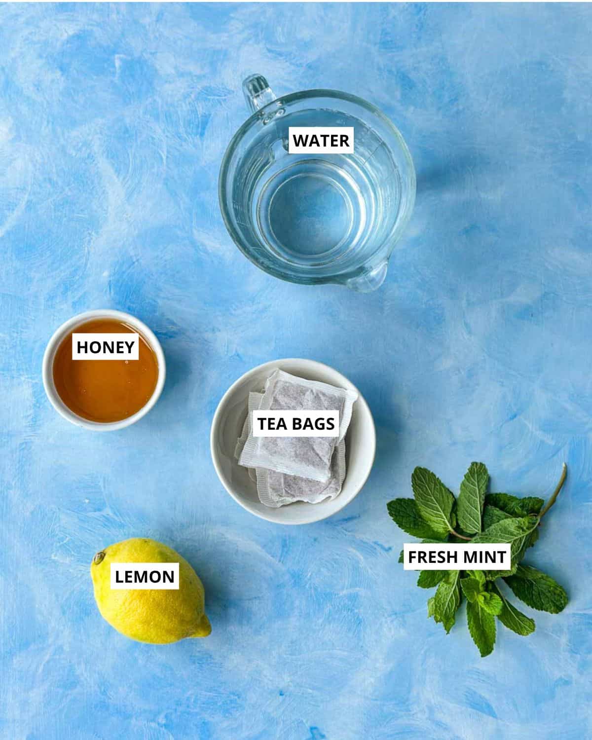 Overhead ingredient shot for lemon iced tea with water, tea bags, honey, lemon, and fresh mint.