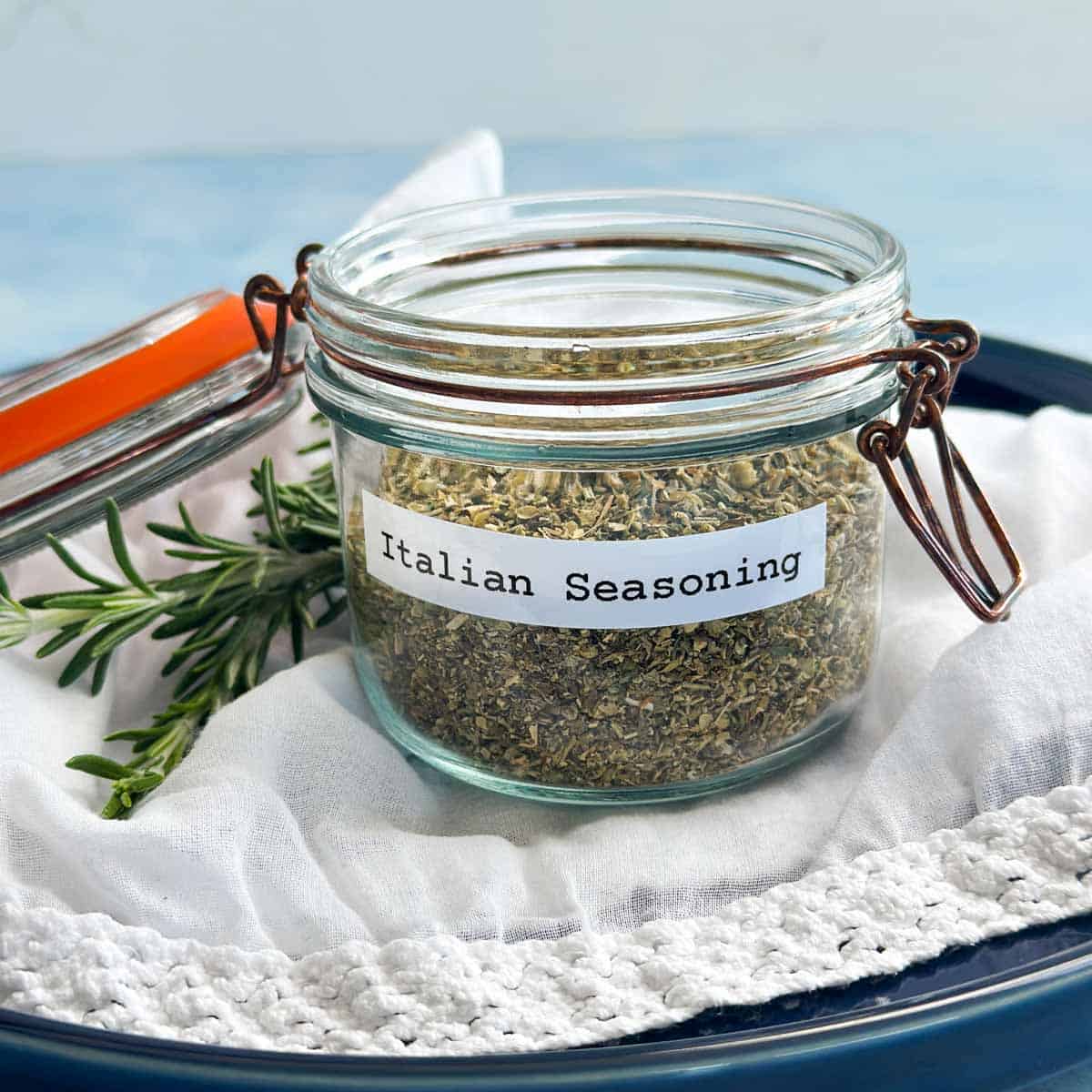 Close-up of homemade Italian seasoning in a glass storage jar with fresh rosemary and white linen styling on a dark blue plate.