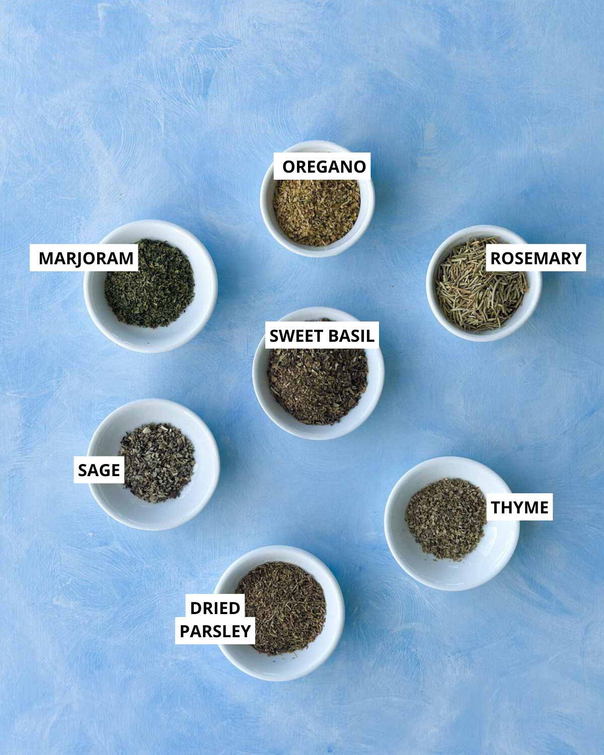 Overhead flat lay of dried herbs used for homemade Italian seasoning including oregano, rosemary, thyme, sage, marjoram, sweet basil, and dried parsley on a pale blue background.
