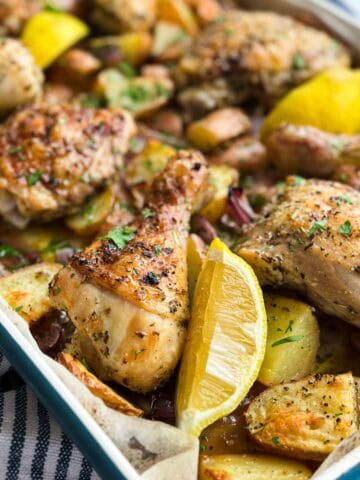Close-up view of golden roasted chicken, tender potatoes, red onion, and bright lemon wedges in a sheet pan dinner.