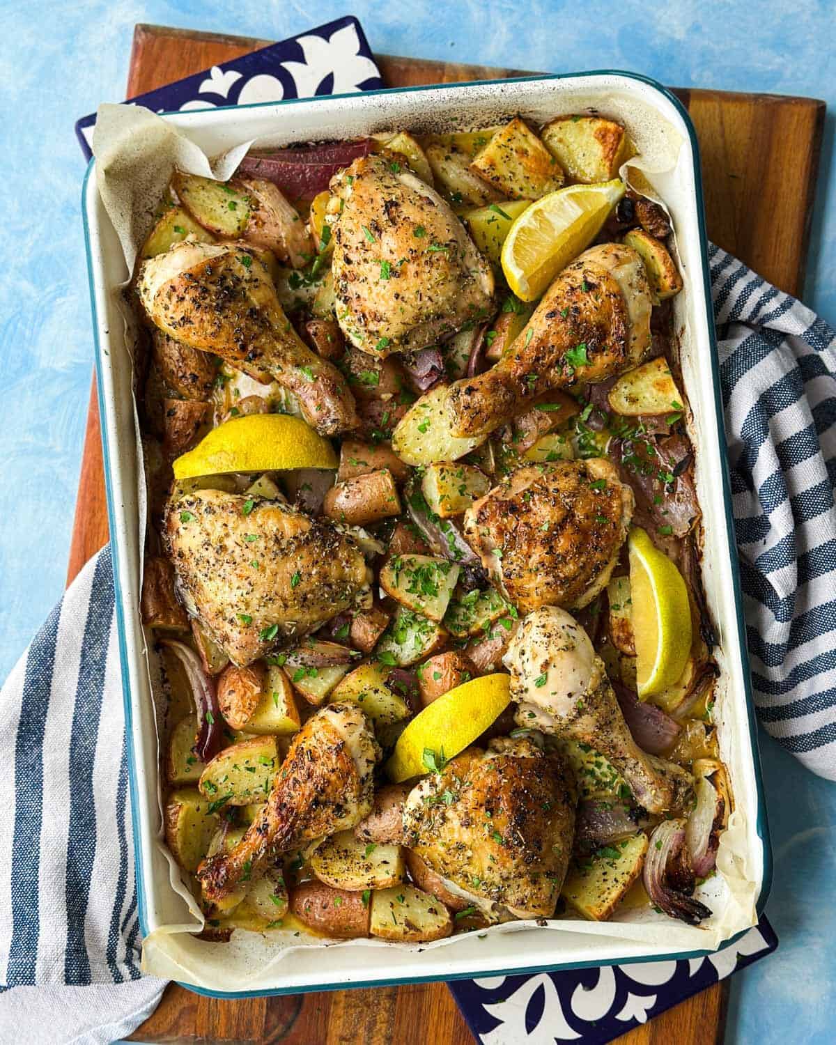 Overhead shot of sheet pan chicken and potatoes with crisp golden skin, roasted red onion, lemon wedges, and parsley.