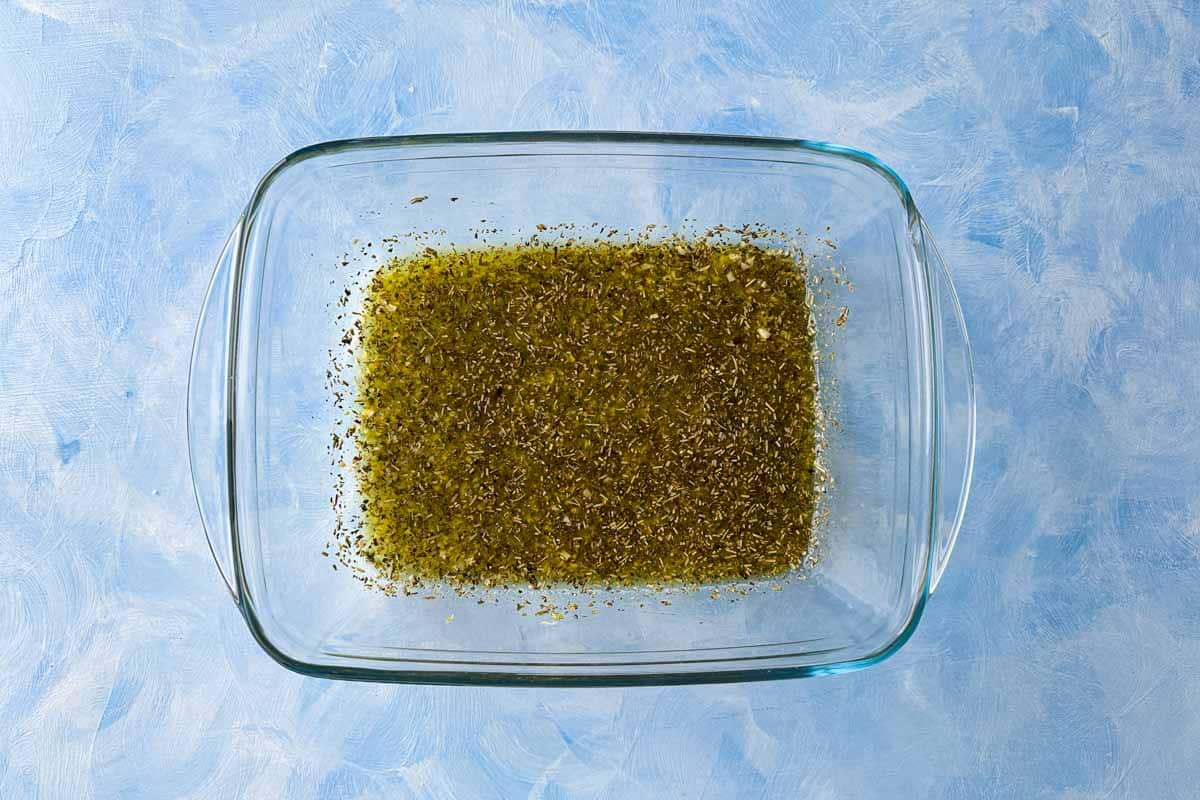 Top-down view of olive oil, garlic, and Italian herbs mixed in a glass baking dish for the chicken marinade.