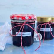 Two jars of homemade mixed berry jam with red twine, gingham lid, and deep berry color on a light blue surface.