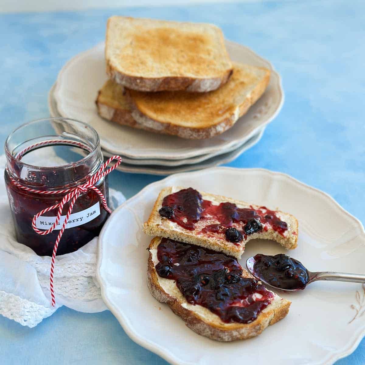 Mixed berry jam served on buttered toast with a spoonful of jam beside the plate and a jar wrapped in red twine.
