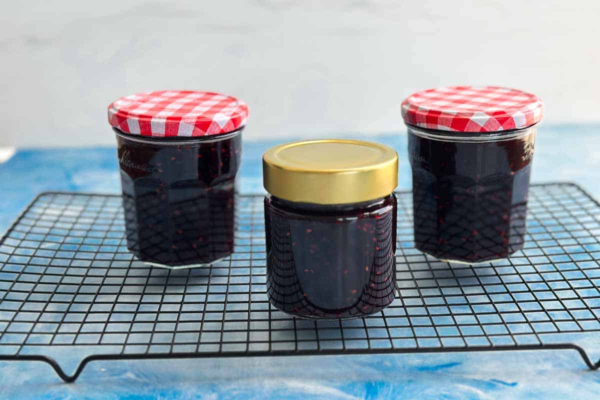 Filled jars of mixed berry jam cooling on a wire rack with red gingham and gold lids