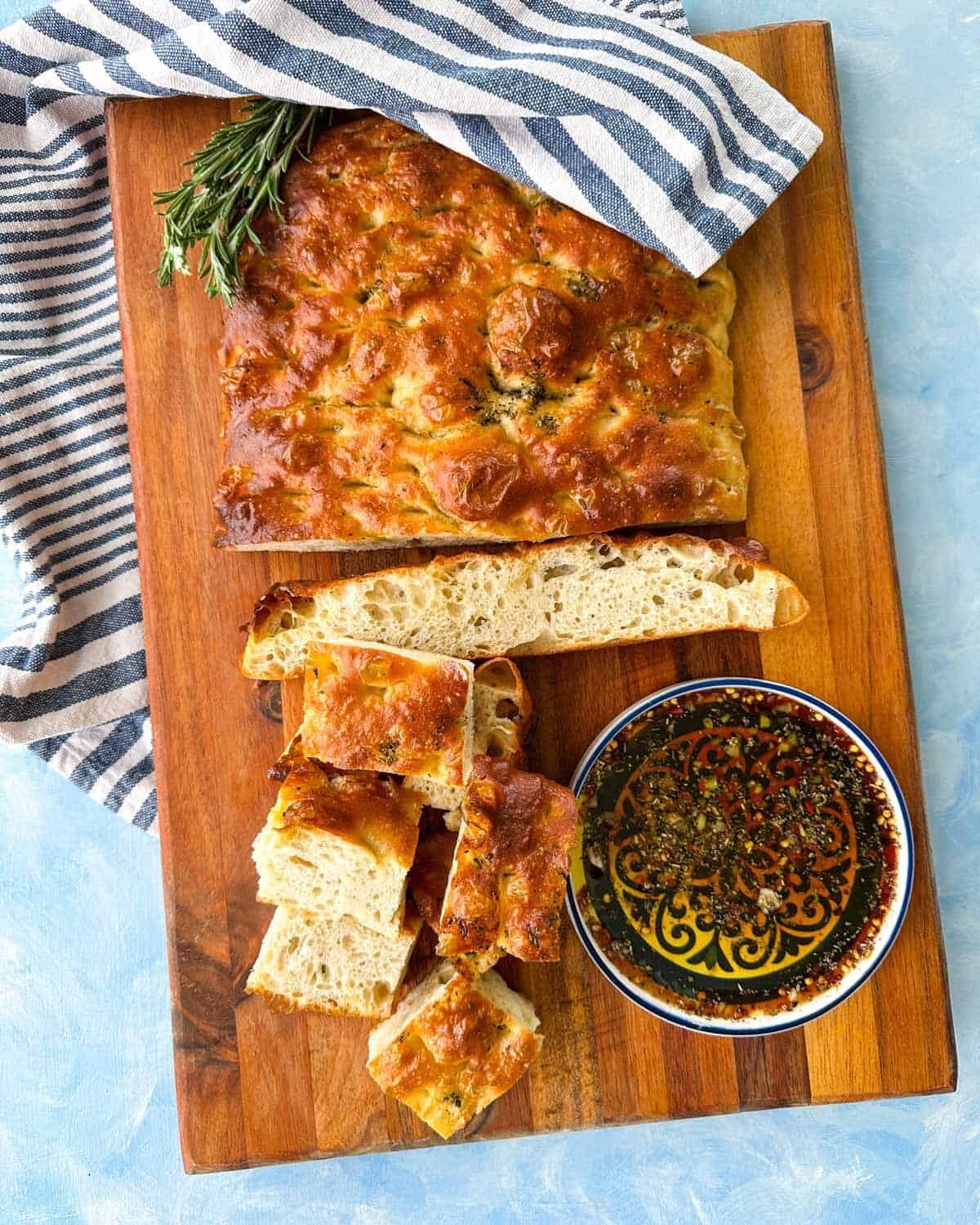 Overhead view of no knead focaccia bread served with olive oil dip, sliced pieces, rosemary, and a striped linen cloth.