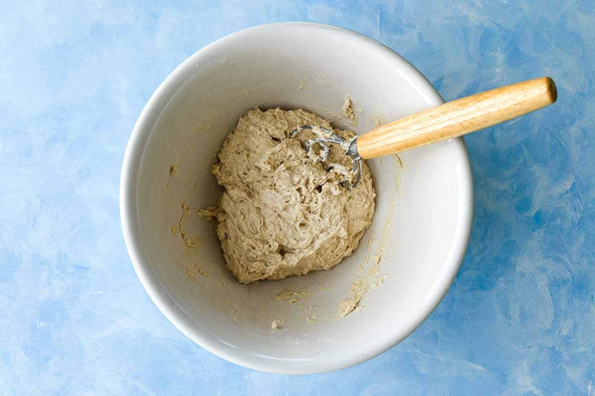 Top-down view of sticky focaccia dough mixed in a white bowl with a Danish dough whisk on a blue work surface.