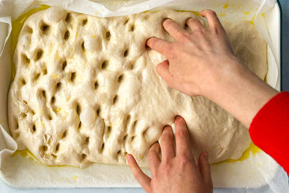 Fingers dimpling risen focaccia dough in an oiled baking dish before adding toppings.