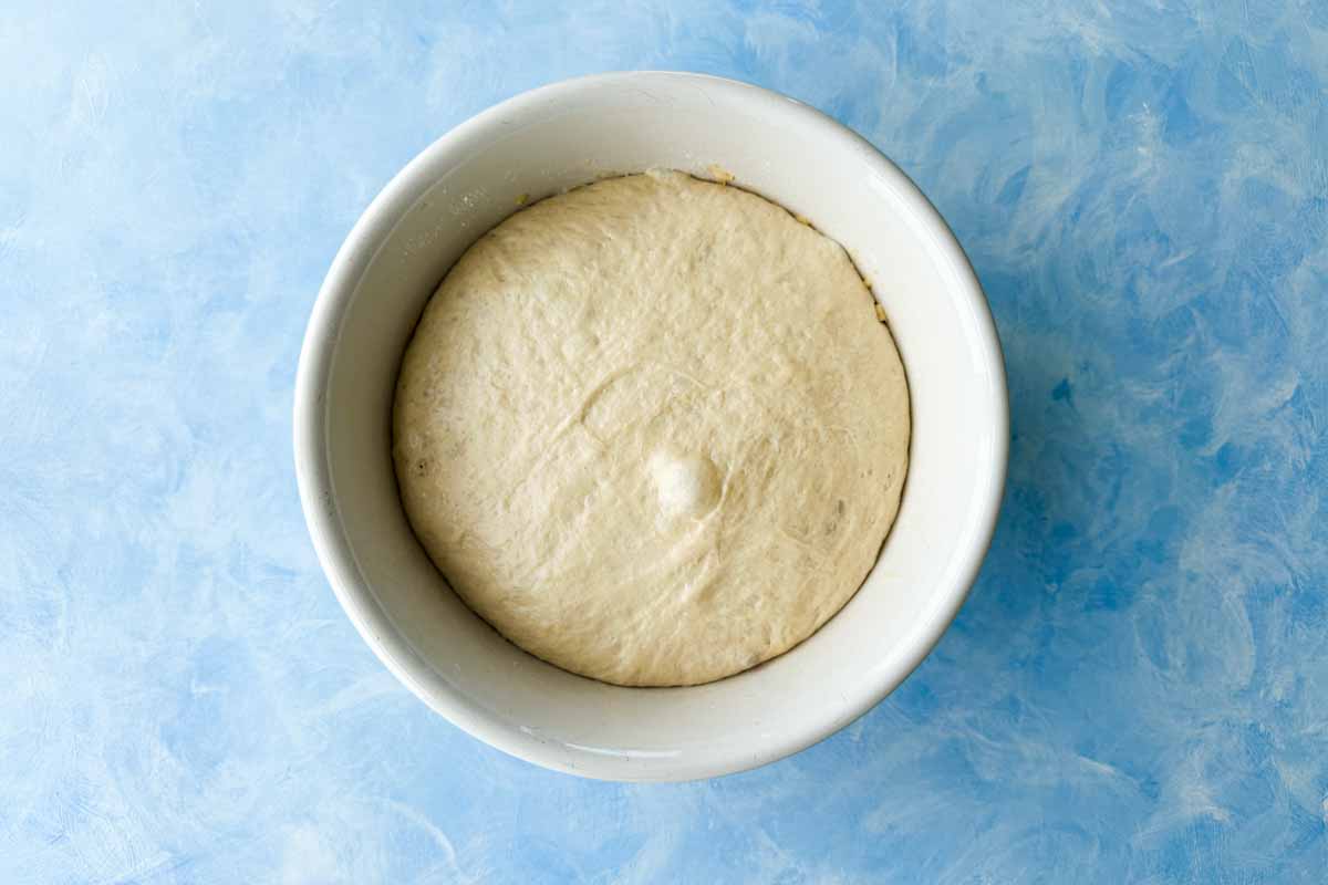 Smooth risen focaccia dough filling a white bowl after resting on a blue work surface.