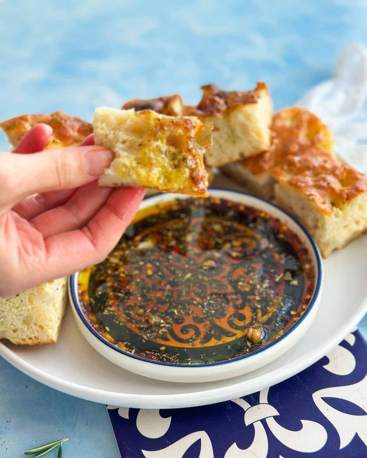 Hand holding a piece of focaccia dipped in olive oil bread dip with garlic, herbs, and balsamic vinegar