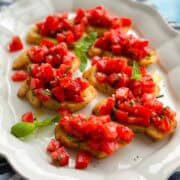Close angled view of fresh tomato bruschetta appetizer featuring toasted crostini topped with ripe tomatoes, basil, garlic, and olive oil, styled on a white platter with soft natural lighting.