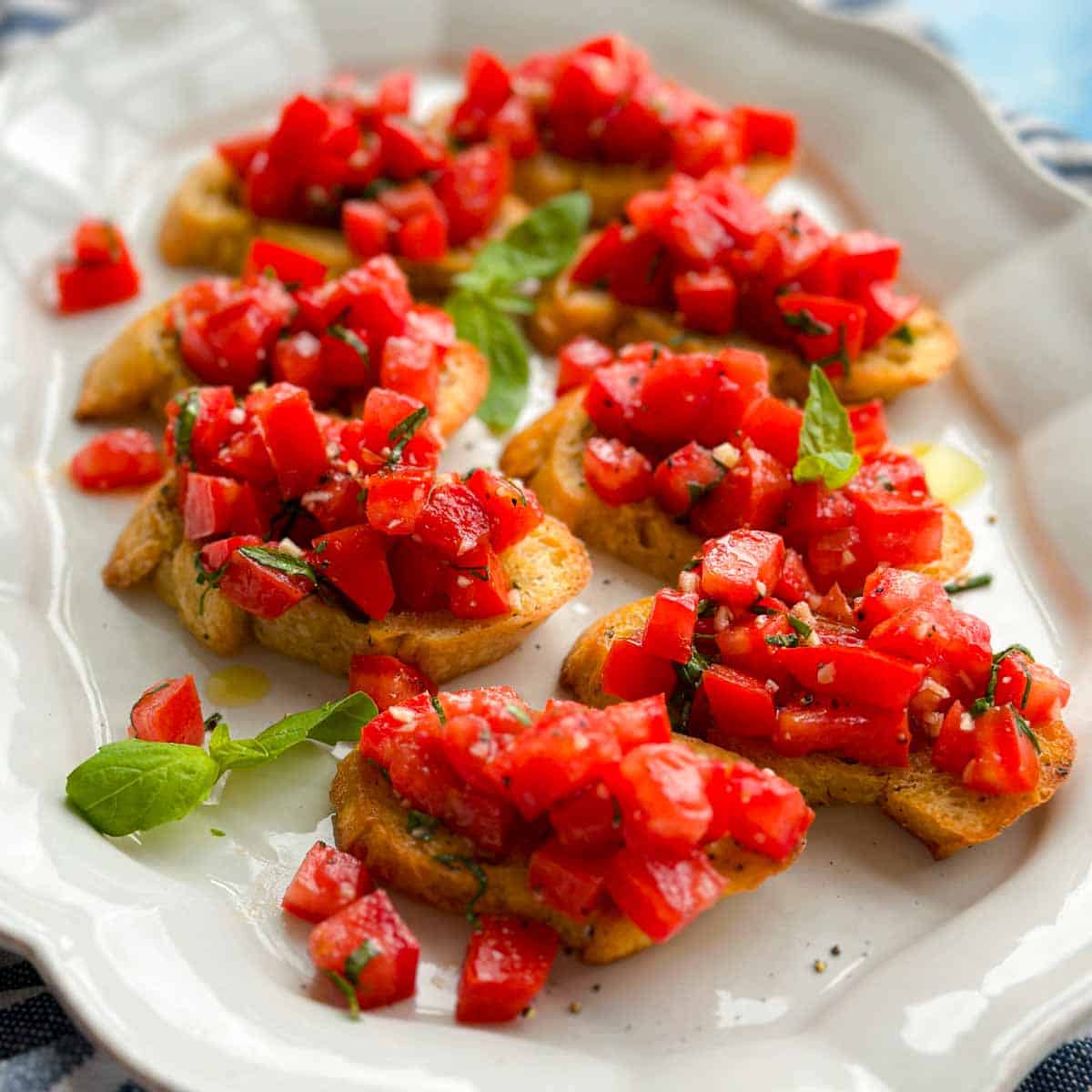 Close angled view of fresh tomato bruschetta appetizer featuring toasted crostini topped with ripe tomatoes, basil, garlic, and olive oil, styled on a white platter with soft natural lighting.