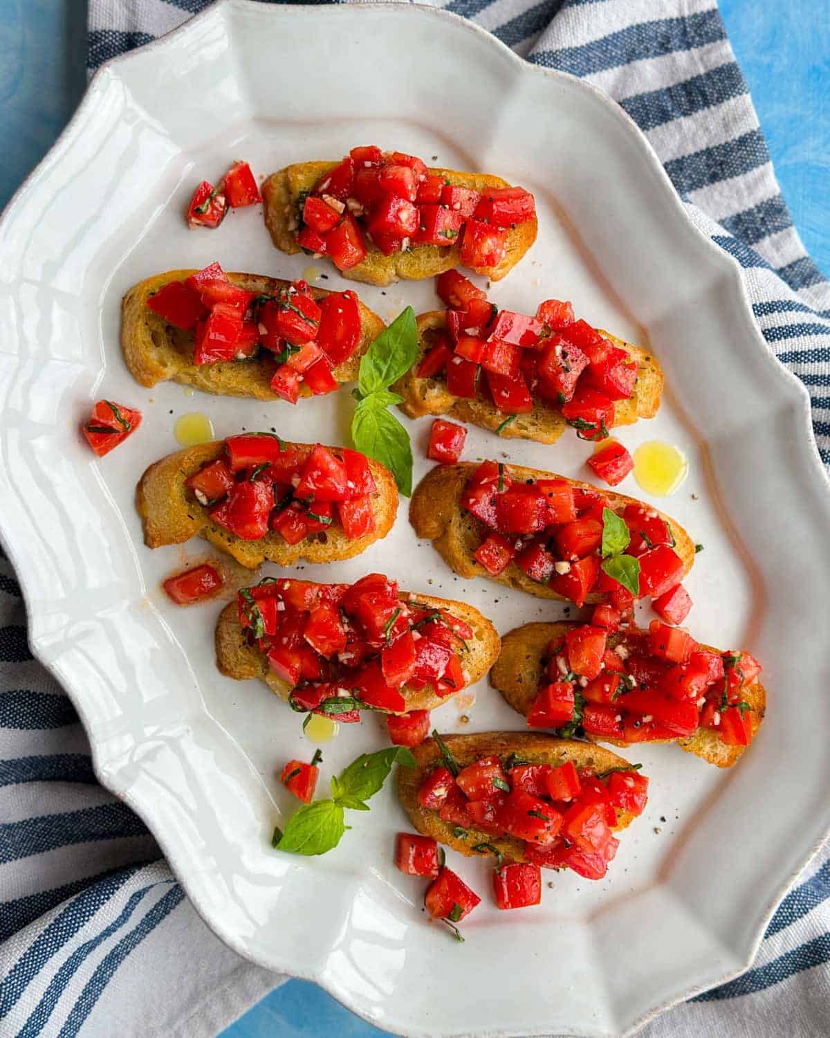 Overhead view of easy tomato bruschetta arranged neatly on a white ceramic platter with toasted baguette slices, fresh basil leaves, diced tomatoes, and olive oil scattered across the plate.