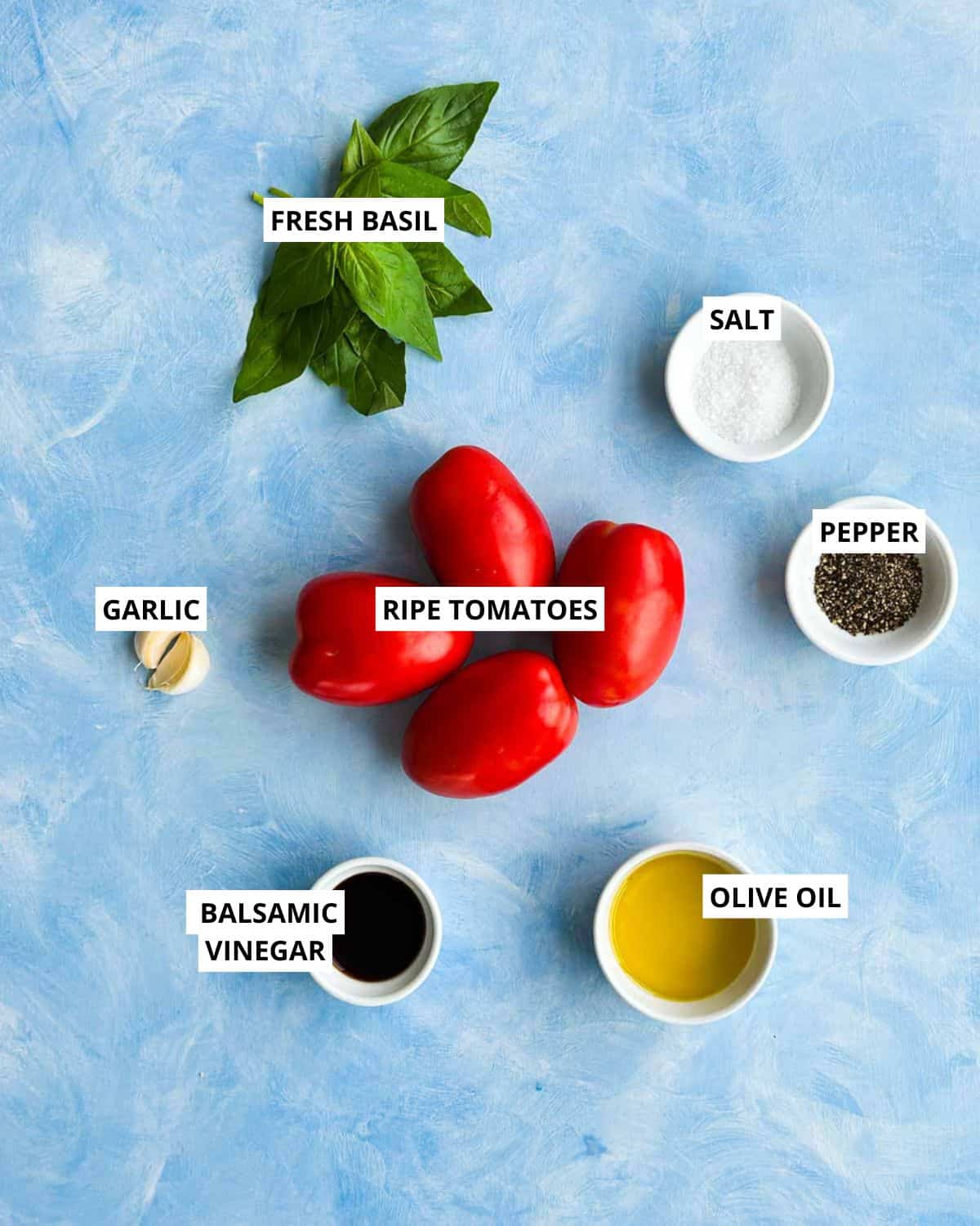 Overhead ingredient image for tomato bruschetta showing ripe tomatoes, fresh basil, garlic, balsamic vinegar, olive oil, salt, and black pepper arranged on a blue textured background with labels
