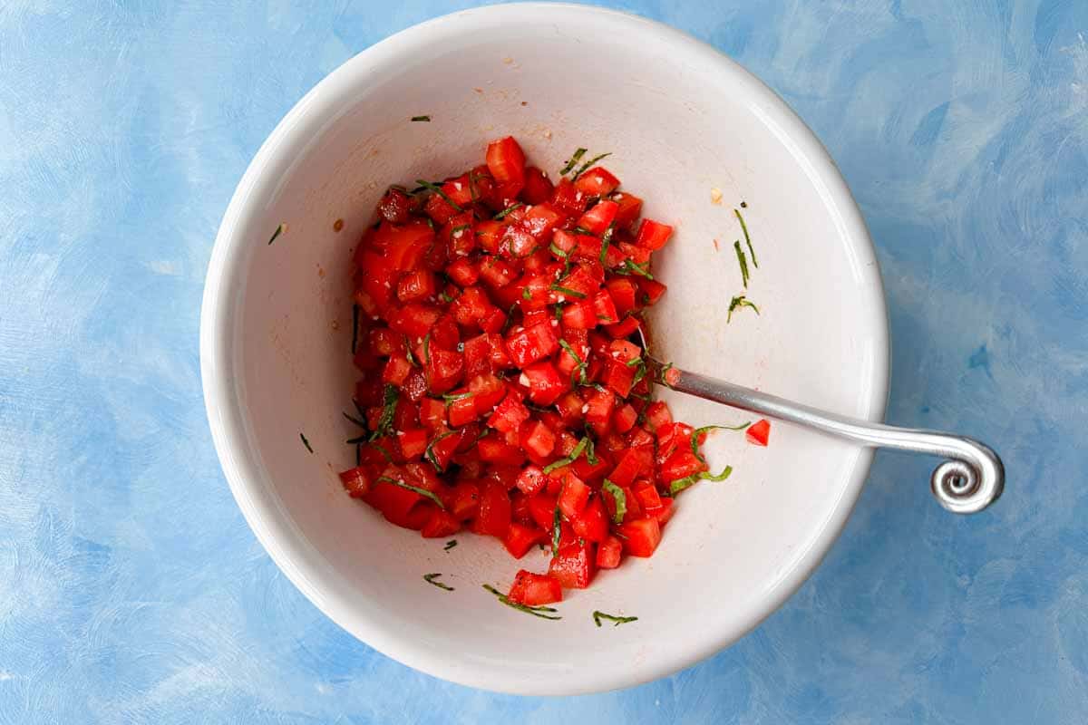 Top view of fresh tomato bruschetta topping mixture with diced tomatoes, basil, garlic, olive oil, and balsamic vinegar mixed in a white bowl with a spoon.