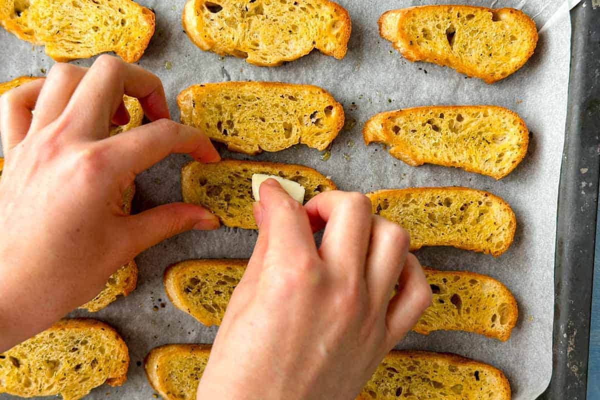 Close-up process image of hands rubbing fresh garlic over warm toasted baguette slices on a baking tray to prepare flavorful bruschetta bread.