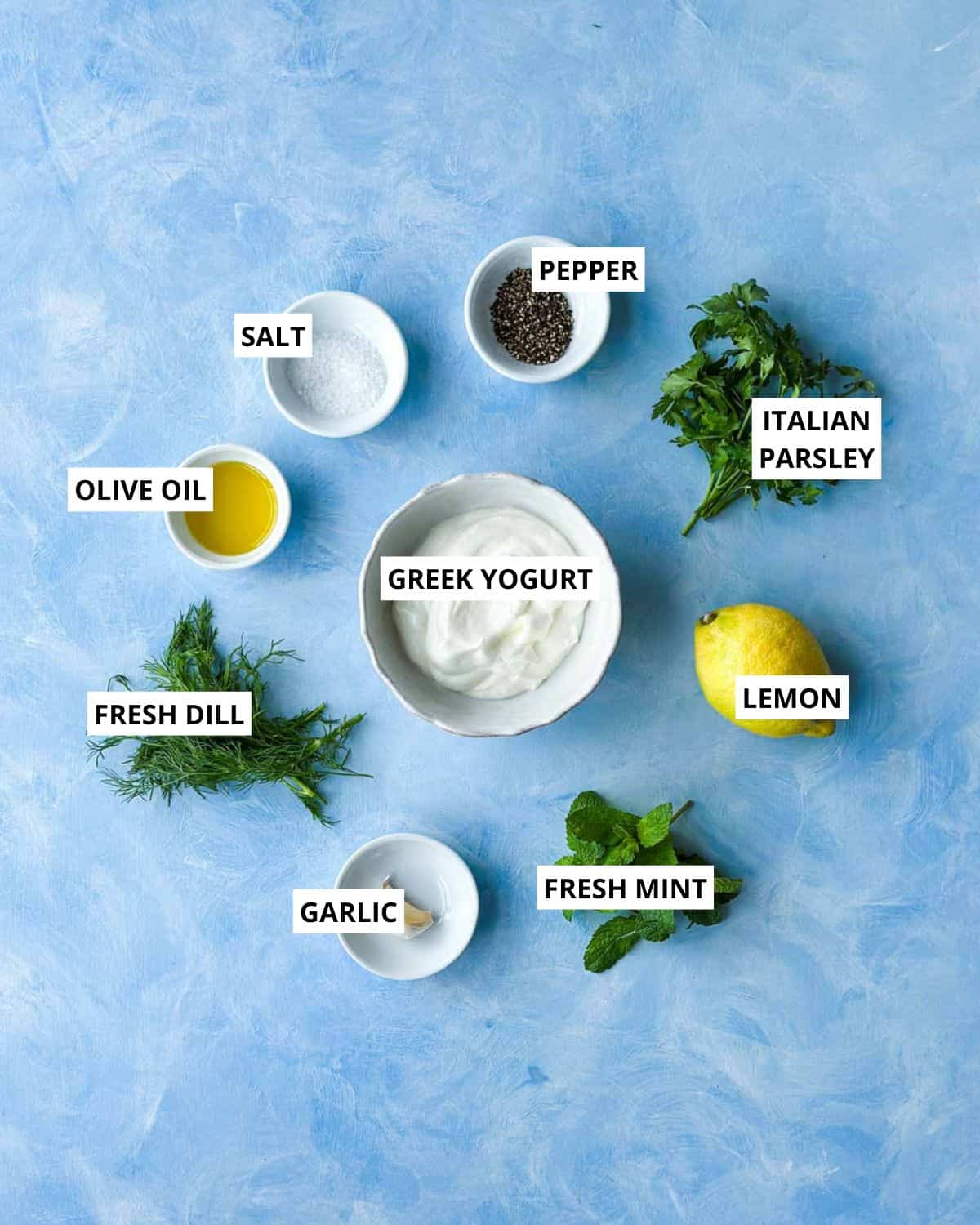 Overhead ingredient layout for creamy herb dressing with Greek yogurt, dill, mint, parsley, garlic, lemon, olive oil, salt, and pepper.