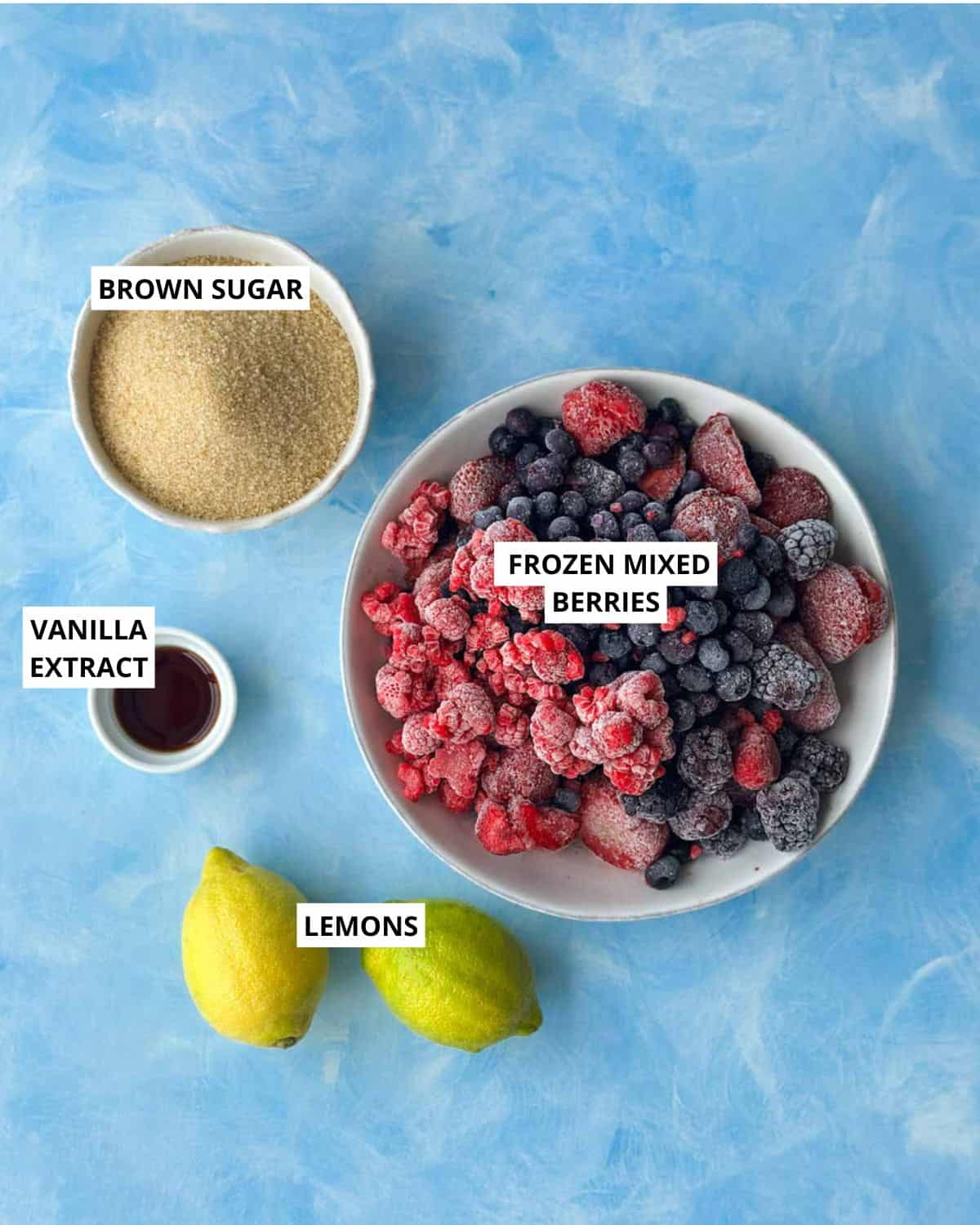 Top-down view of frozen mixed berries, brown sugar, lemons and vanilla extract for homemade jam.