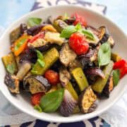 Close-up of roasted vegetables in a white bowl with eggplant, zucchini, cherry tomatoes, peppers, and fresh basil leaves.
