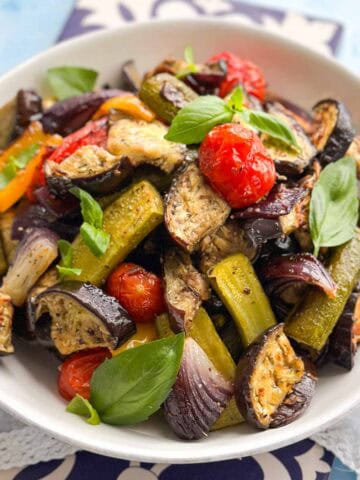 Close-up of roasted vegetables in a white bowl with eggplant, zucchini, cherry tomatoes, peppers, and fresh basil leaves.