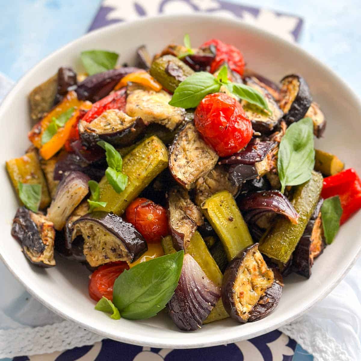 Close-up of roasted vegetables in a white bowl with eggplant, zucchini, cherry tomatoes, peppers, and fresh basil leaves.