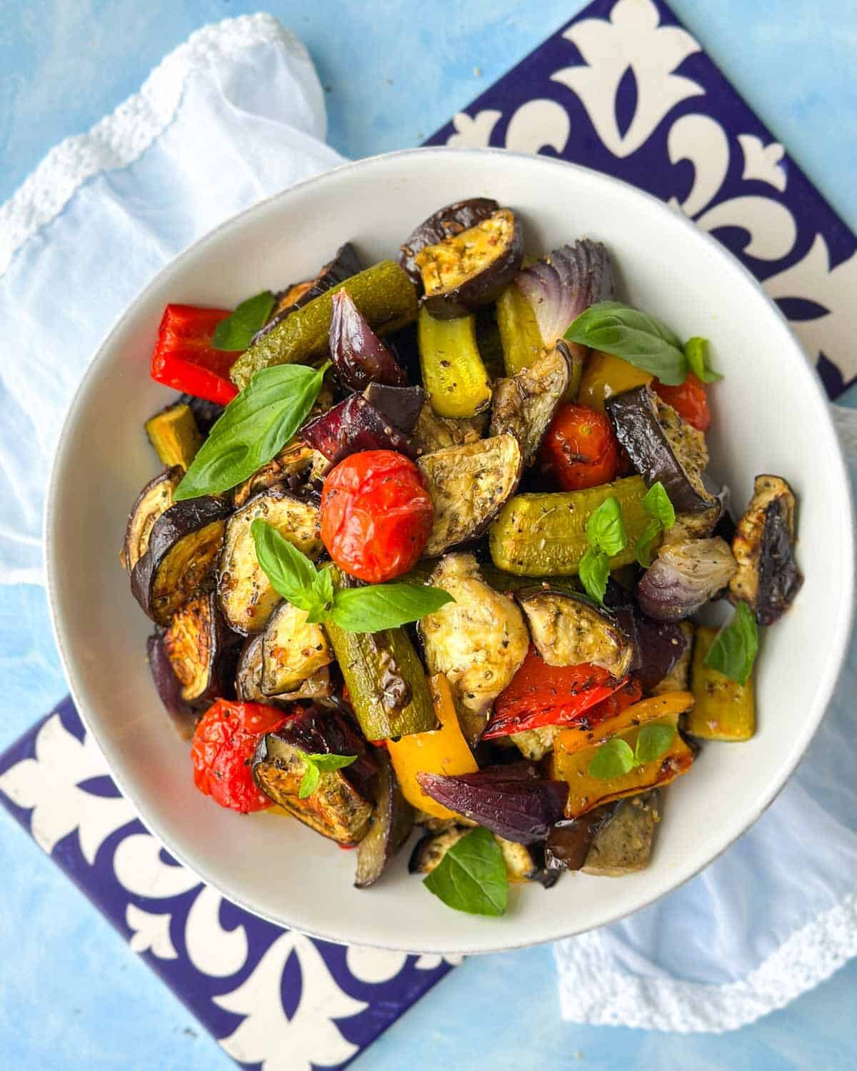 Overhead view of Mediterranean roasted vegetables in a white bowl with basil, eggplant, zucchini, red onion, and peppers.