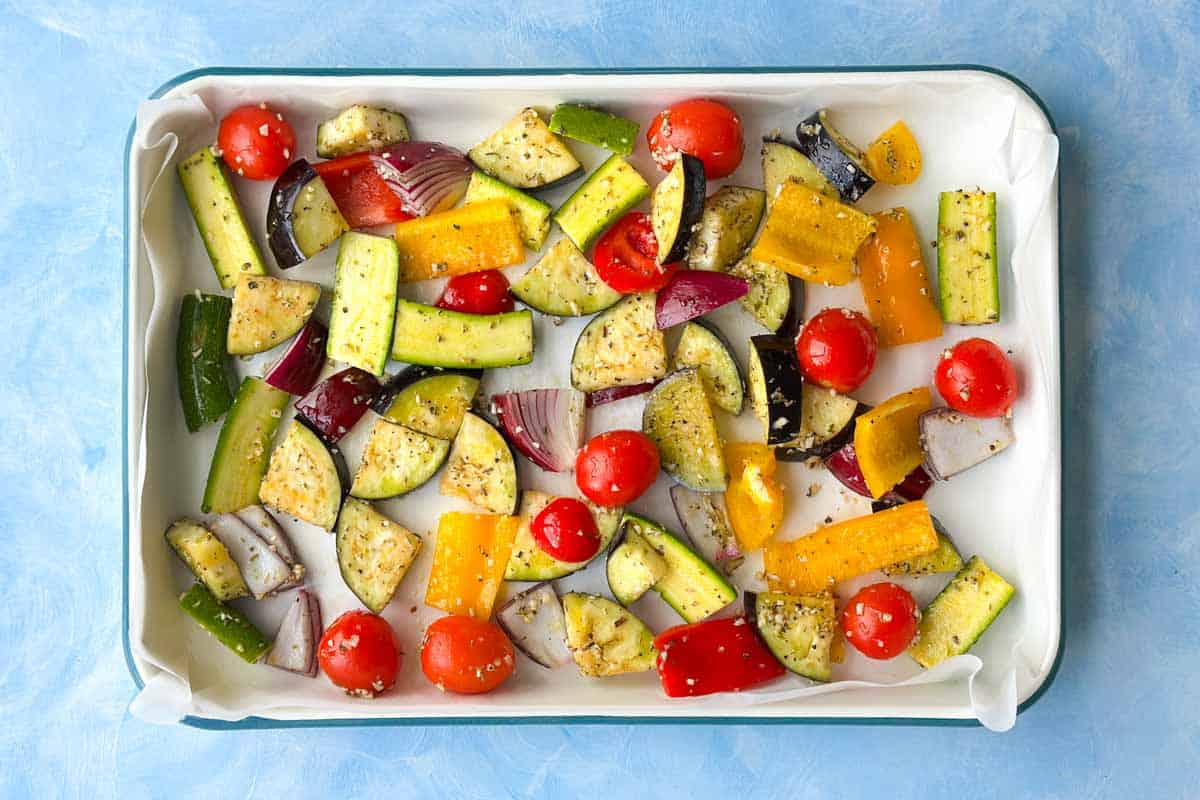 Raw Mediterranean roasted vegetables spread in a baking dish before roasting, including eggplant, zucchini, peppers, onion, and tomatoes.