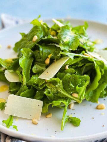 Side view of rocket salad with crisp arugula, shaved Parmesan, pine nuts, and lemon zest on a white plate.