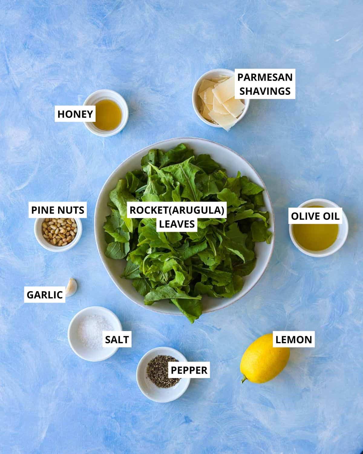 Overhead ingredient shot for rocket salad with arugula leaves, Parmesan shavings, pine nuts, lemon, garlic, olive oil, honey, salt, and pepper.