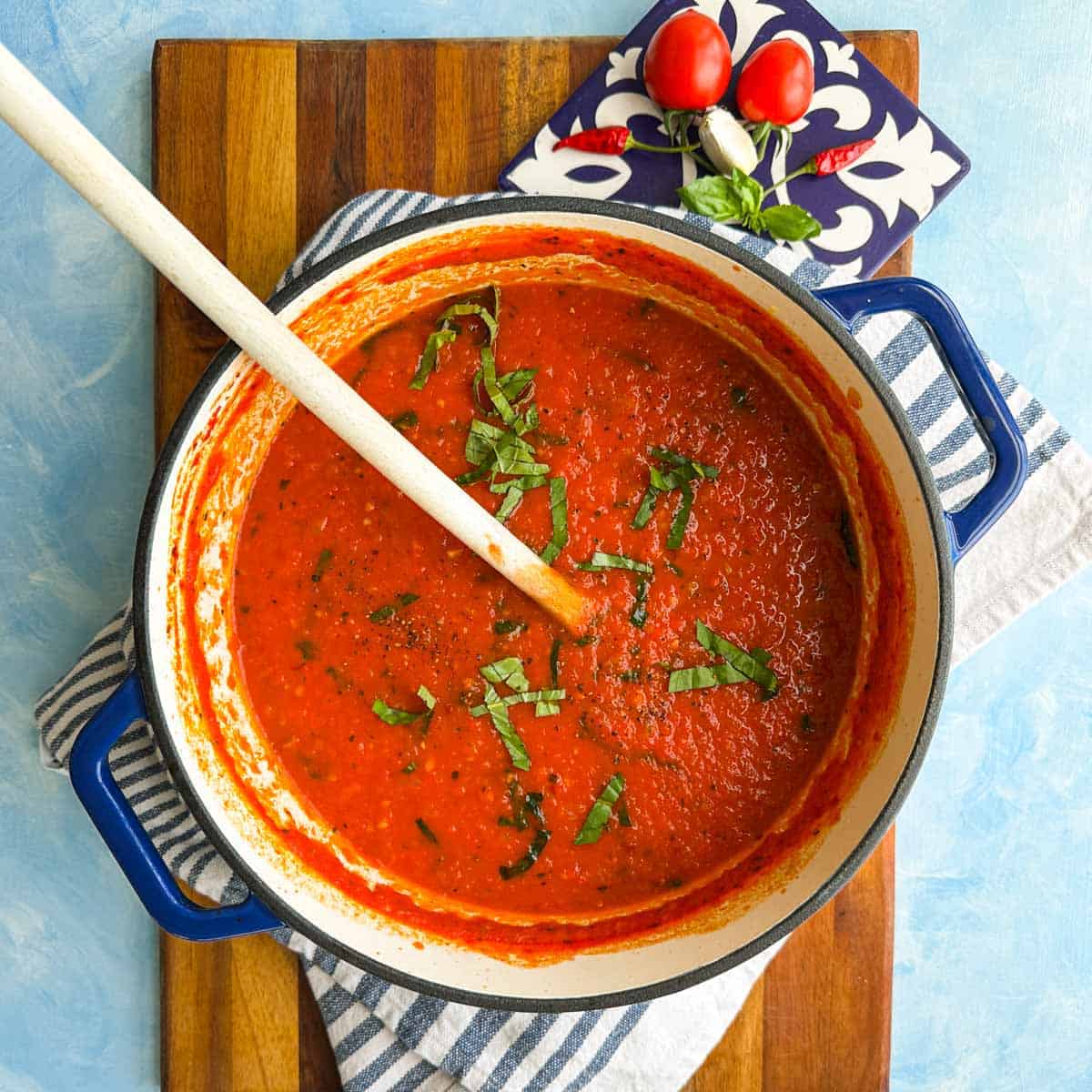 Spicy tomato pasta sauce in a blue Dutch oven with basil, tomatoes, garlic, and chilies styled beside the pot.