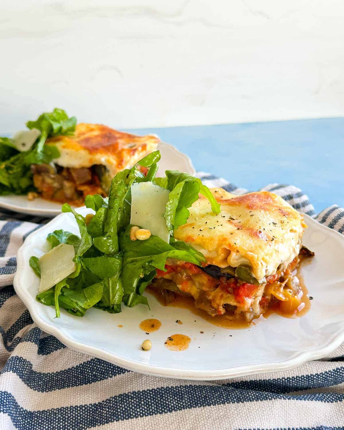 Side view of a plated serving of Mediterranean vegetable lasagna with fresh salad and Parmesan, with another plate softly blurred behind.