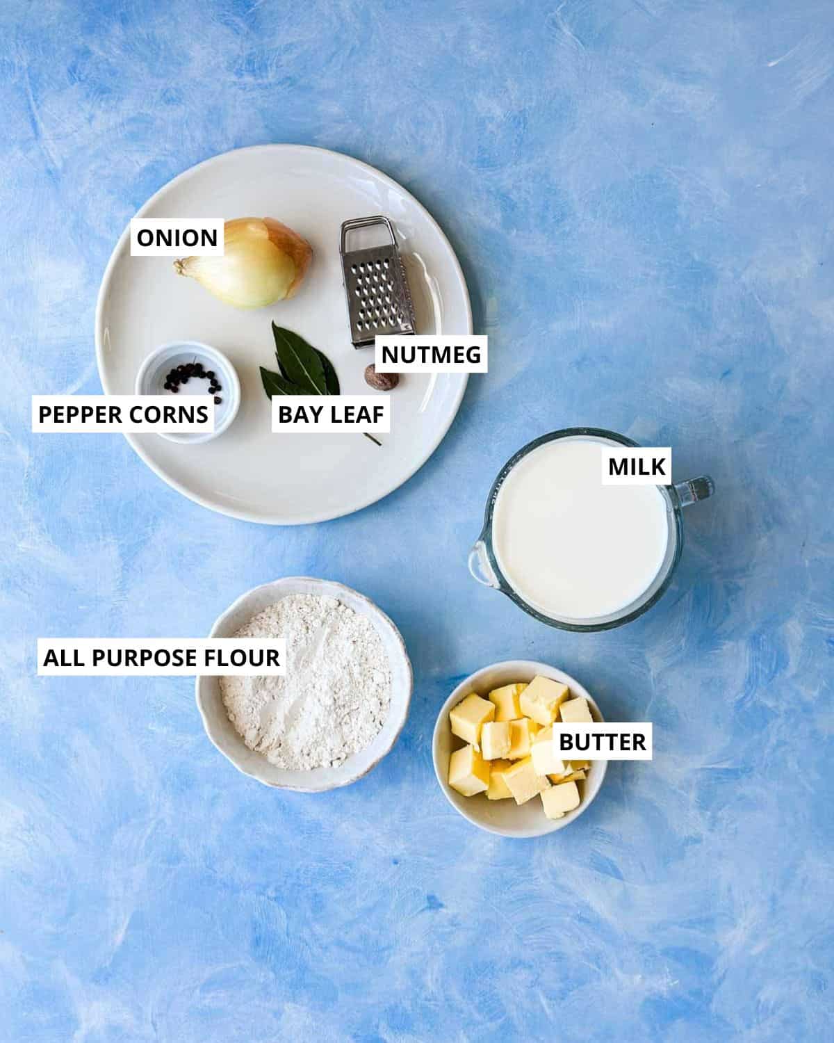 Top-down view of béchamel ingredients for Mediterranean vegetable lasagna with milk, butter, flour, onion, bay leaf, nutmeg, and peppercorns.