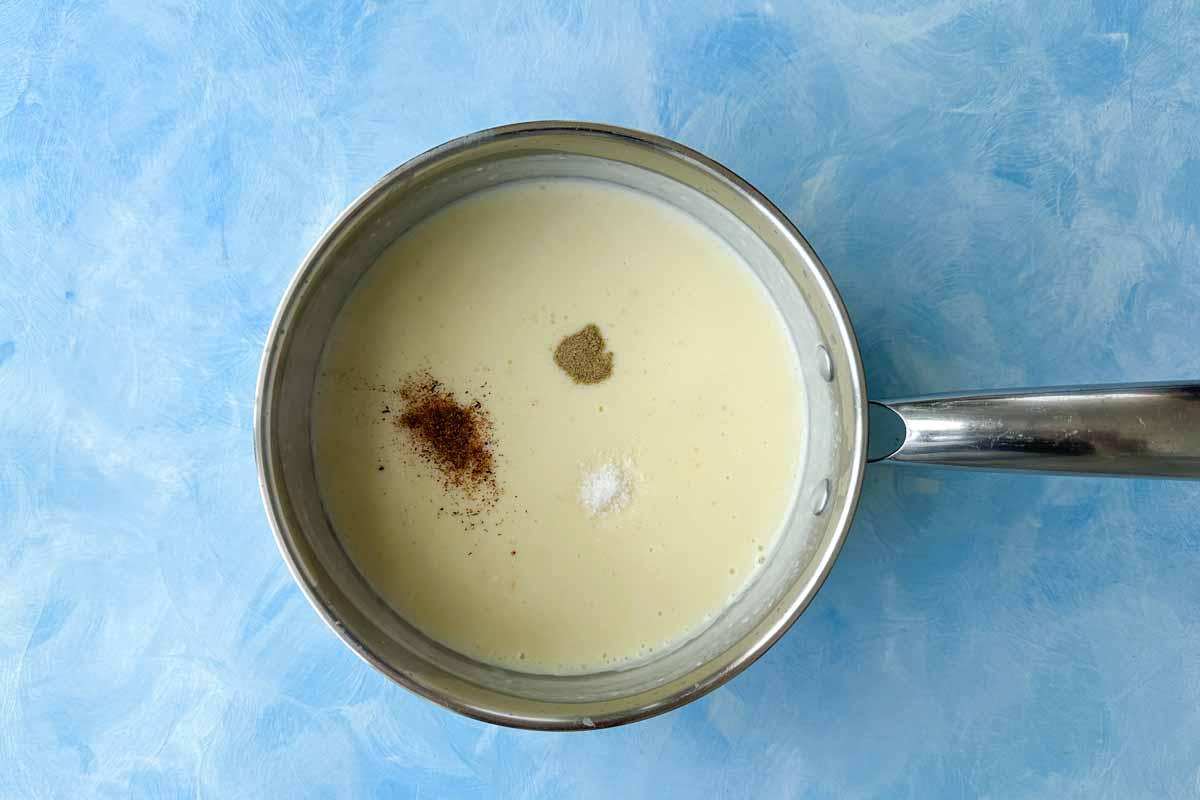 Top-down view of béchamel sauce seasoned with nutmeg, salt, and pepper in a saucepan.