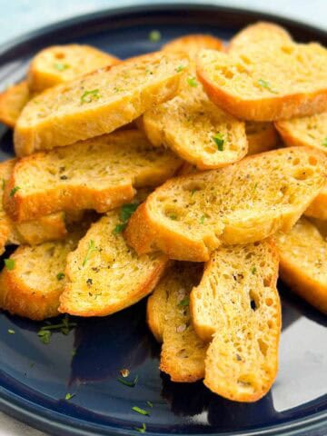 Close-up view of crunchy crostini stacked on a dark blue plate with herbs scattered over the toasted bread slices.