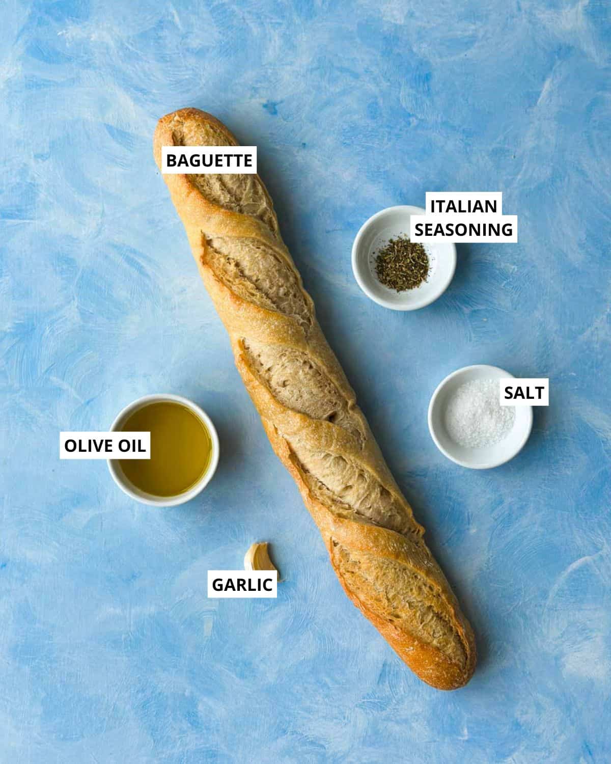 Overhead ingredient layout for homemade crostini with baguette, olive oil, garlic, Italian seasoning, and salt.