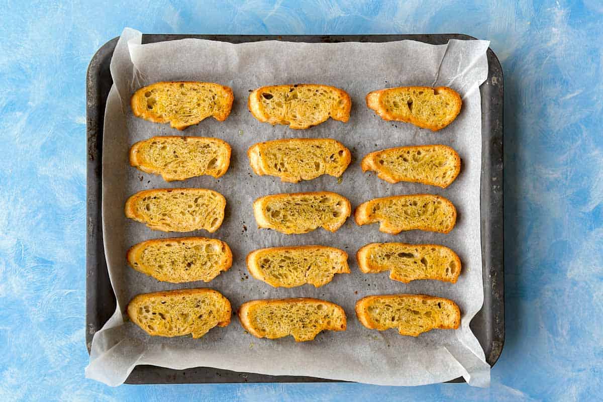 op-down view of baked crostini on a lined baking tray, toasted until golden and crisp.