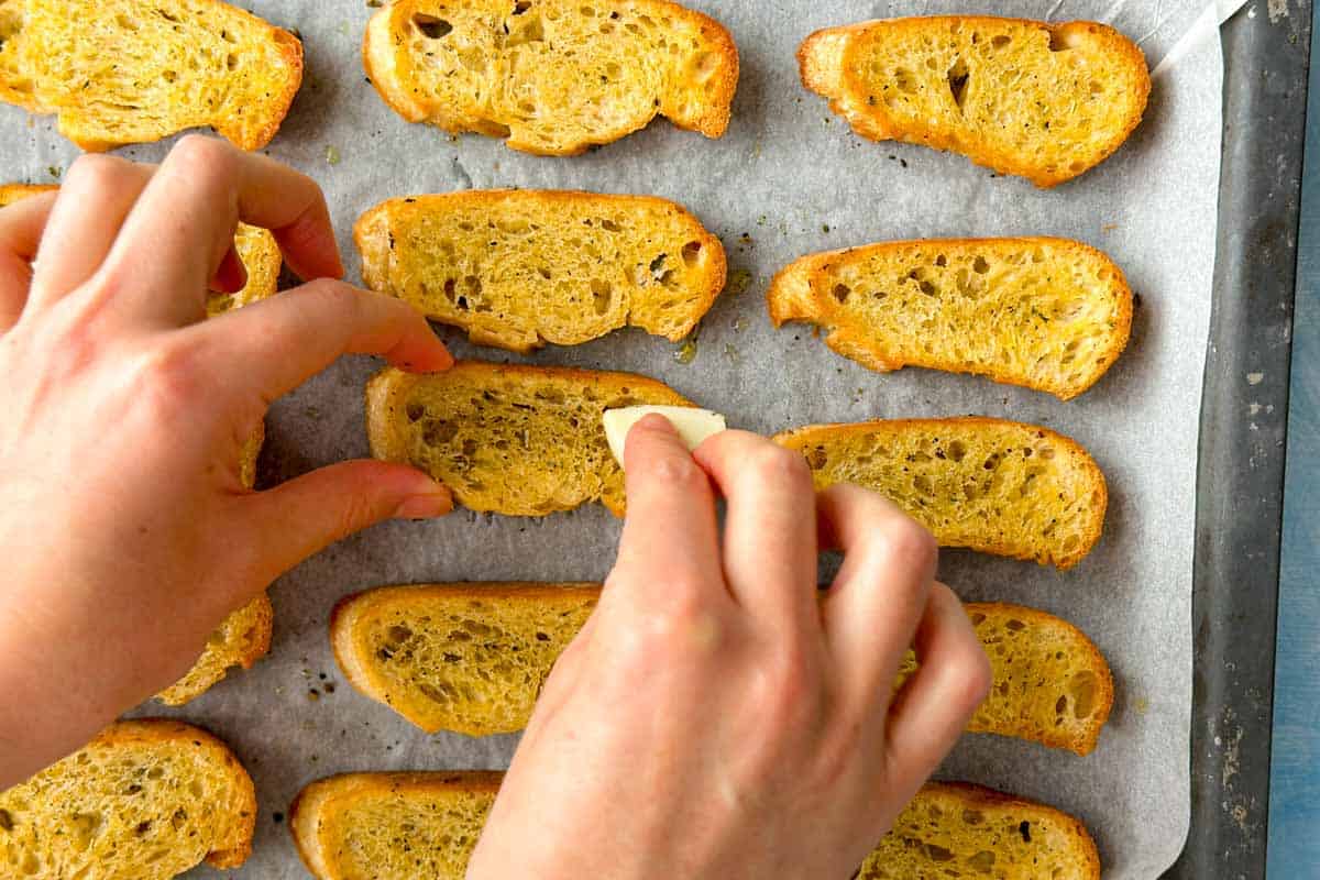 Hands rubbing garlic over warm crostini on a baking tray to add flavor before serving.