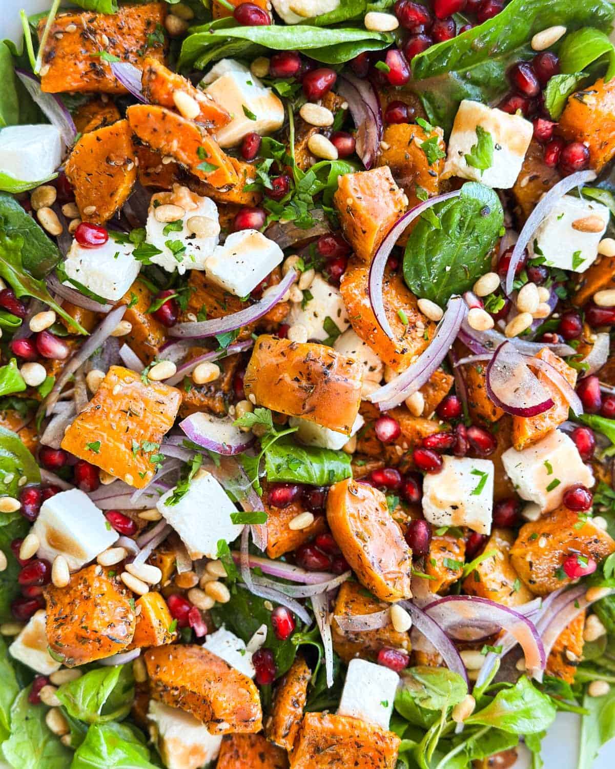 Horizontal image of roasted butternut squash salad with feta, pomegranate arils, pine nuts, red onion, and greens.
