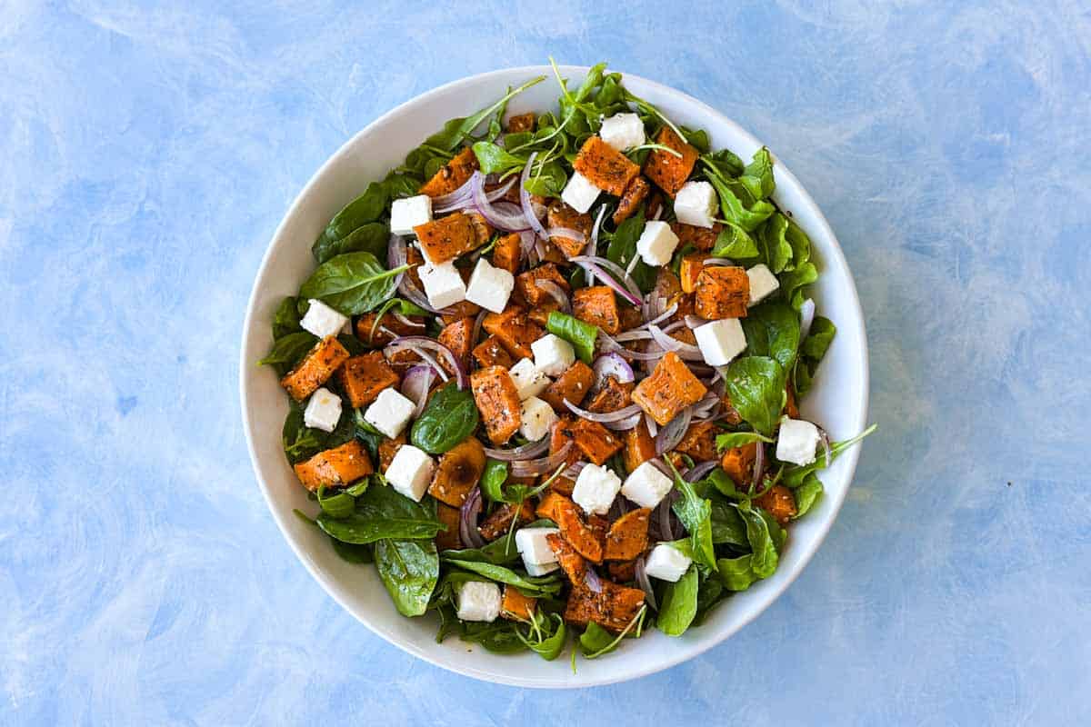 Roasted butternut squash salad being assembled with greens, squash, feta, and red onion in a white bowl.