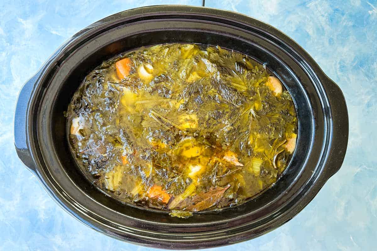 Top view of slow cooker beef stock simmered with beef bones, celery, onion, carrots, garlic, thyme, and bay leaves.