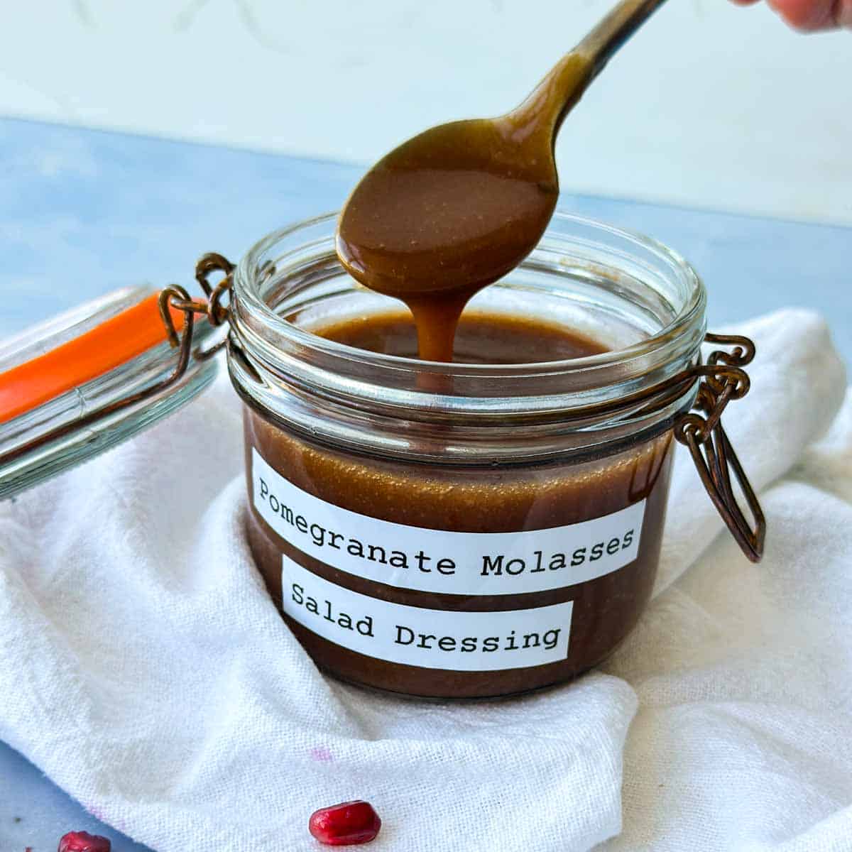 Spoon lifting thick pomegranate molasses dressing from a labeled glass jar on a white cloth.